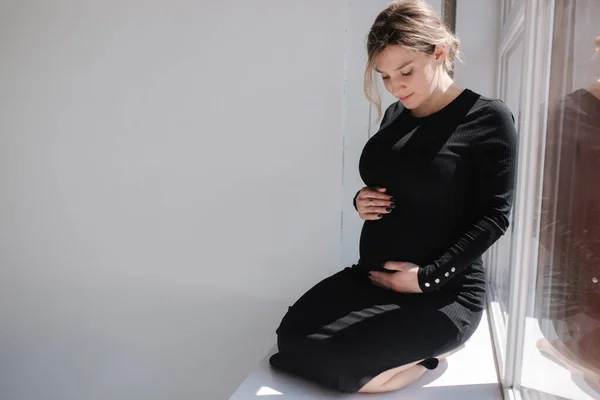 Mulher grávida em vestido preto senta-se no peitoril da janela e colocar as mãos na barriga. Futura mãe está esperando pelo bebê — Fotografia de Stock