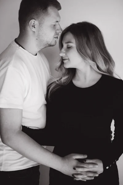 Mujer embarazada con su encantador marido abrazarse y poner la mano en el vientre. Fotosesión de estudio. Foto de fondo en blanco y negro. Hombre besar a su esposa en la frente — Foto de Stock