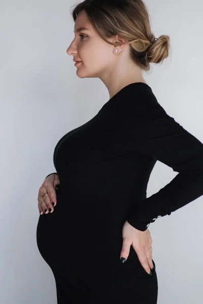 A mulher grávida elegante em dragas de maternidade pretas bonitas põe a mão na barriga no estúdio. Fundo branco — Fotografia de Stock