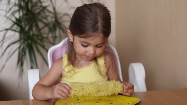 Cute little girl ryb jedzenia kukurydzy i wyciera jej usta. Urocze dziecko w żółtej koszuli stawia huśtawkę kukurydzy na żółtym talerzu. Letni nastrój w domu — Wideo stockowe