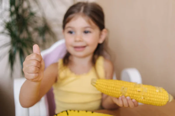 Happy little girl hold fresh corn in hand and smile. Female kid thumbs up. Summer mood at home. Focus on hand — Stock Photo, Image