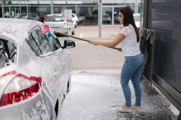 Çekici genç bir kadın arabasını şampuan ve fırçayla yıkıyor. Kadın oto yıkama servisinde köpük ve suyla araba yıkar. — Stok fotoğraf