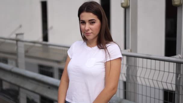 Menina jovem atraente em branco t-shirt stand na frente do shopping no estacionamento ao ar livre. Mulher a olhar para a câmara. Movimento lento — Vídeo de Stock