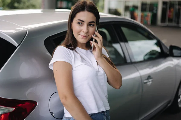 Attraktiv ung kvinna använder sin telefon och lutade sig mot sin bil på bensinstationen. Närbild av kvinna väntar bensinstation arbetare — Stockfoto