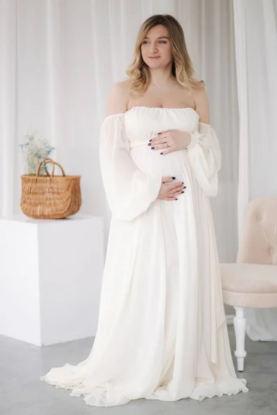 Beautiful pregnant woman in elegant white dress posing to photographer in studio. Background of white tulle — Zdjęcie stockowe