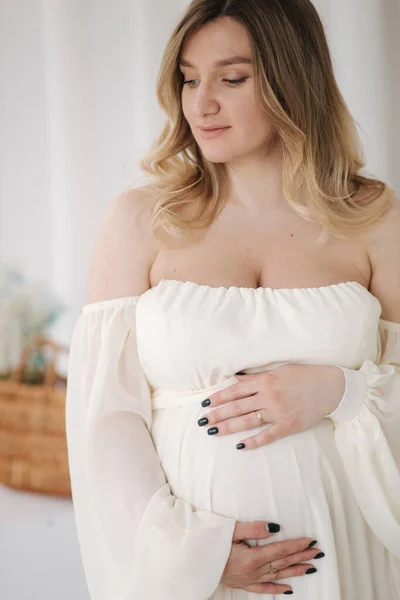 Красива вагітна жінка в елегантній білій сукні позує фотографу в студії. Фон з білого тюлю — стокове фото