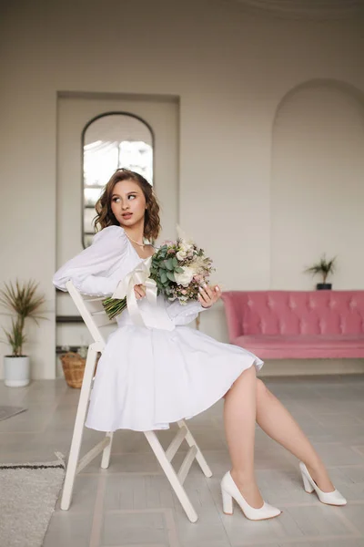 Élégante mariée s'assoit sur le tabouret et tient le bouquet à la main. Photosession de mode en studio — Photo