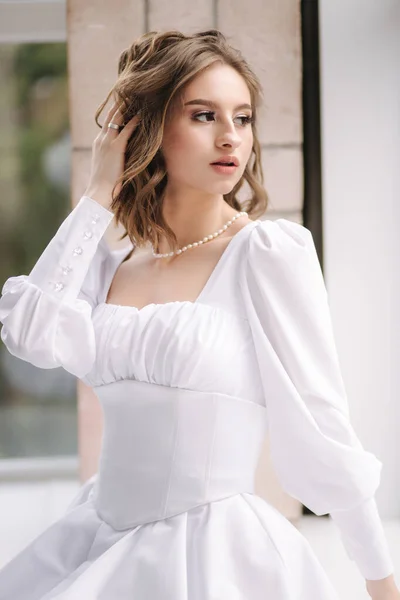 Elegant bride sitting on windowsill and put her hand on hair. Woman in white dress looking into window. Brides morning concept — Stock Photo, Image