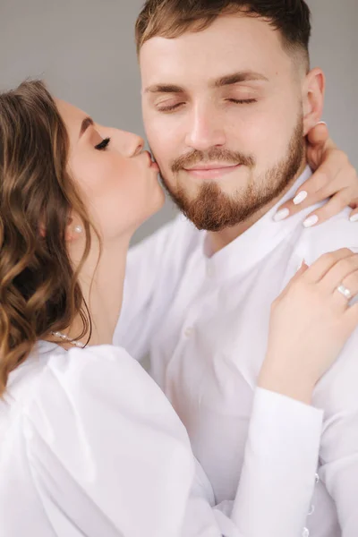 Novio con su adorable novia abrazándose y besándose en la mejilla. Feliz pareja de boda frente a la pared gris. Fotosesión Stuio — Foto de Stock