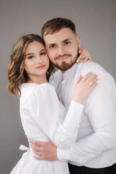 Der Bräutigam und die hübsche Braut umarmen sich und lächeln in die Kamera. Glückliches Hochzeitspaar vor grauer Wand. Stuio Fotosession — Stockfoto