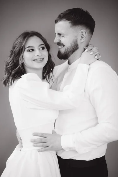 Groom with her lovely bride hug each other and make faces. Happy wedding couple in front of grey wall. Stuio photosession. Black and white photo — Stock Photo, Image