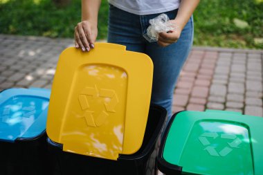 Parkta geri dönüşüm kutusuna plastik poşet atan bir kadın. Plastik kutularda farklı renkler var. Yeşil, mavi ve sarı
