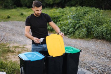 Arka bahçedeki geri dönüşüm kutusuna plastik poşet atan adam. Plastik kutularda farklı renkler var. Yeşil, mavi ve sarı