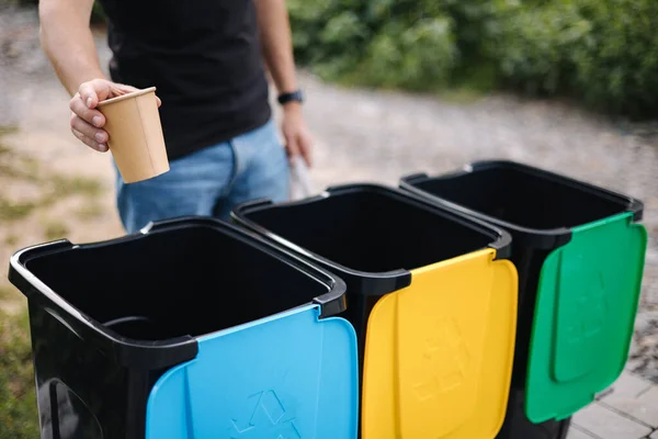 Primo piano della mano umana che getta una tazza di cartone vuota dal cappuccino nel cestino del riciclaggio. Cestini per il riciclaggio degli alberi all'aperto — Foto Stock