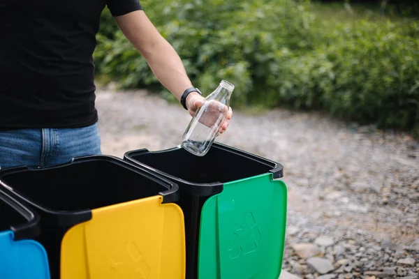 Primo piano della mano umana che lancia una bottiglia di vetro vuota nel cestino del riciclaggio. Cestini per il riciclaggio degli alberi all'aperto — Foto Stock