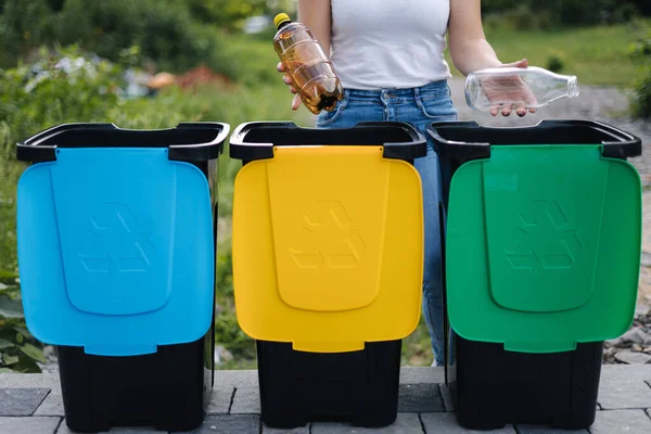 Die mittlere Auswahl an Weibchen hält zwei verschiedene Flaschen, Plastik und Glas und wird in verschiedene Recyclingbehälter geworfen. Großaufnahme einer Frau, die neben blauer, gelber und grüner Tonne steht — Stockfoto