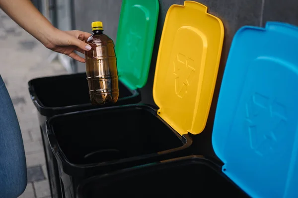 Primo piano della bottiglia di plastica femminile gettata nel cestino del riciclaggio. Diverso colore dei bidoni di riciclaggio all'aperto. Vista laterale della donna mettere spazzatura in contenitore — Foto Stock