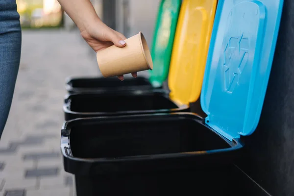 Primo piano di femmina lancio tazza di cartone nel cestino di riciclaggio. Diverso colore dei bidoni di riciclaggio all'aperto. Vista laterale della donna mettere spazzatura in contenitore — Foto Stock