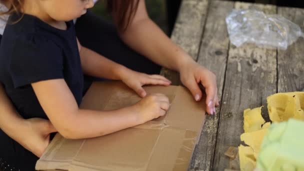 Közelkép egy kislányról az anyjával, aki kartont készít az újrahasznosításhoz. Családi szemétszedés otthon. Az újrafeldolgozás fogalma — Stock videók
