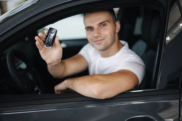 Mannen sitter i sin nya bil i bilens utställningsrum. Porträtt av stilig man i bilen hålla nycklar i händerna — Stockfoto