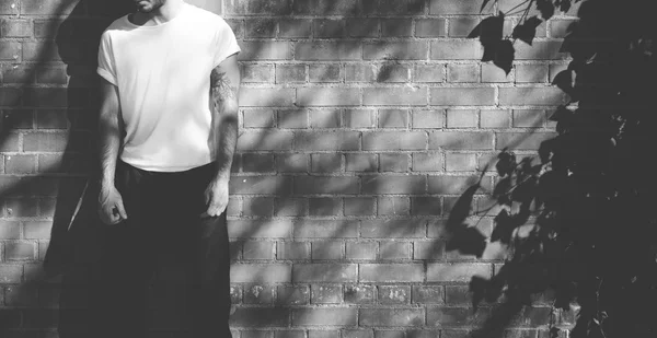 Foto de homem barbudo com tatuagem vestindo tshirt branca em branco e jeans pretos. Parede de tijolos fundo. Ampla maquete, sombras, preto-branco — Fotografia de Stock