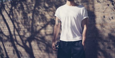 Fotoğraf genç ciddi adam dövme boş beyaz tshirt giyiyor. Bir tuğla duvar önünde duruyor. Şehir sokak arka plan. Yatay mockup.