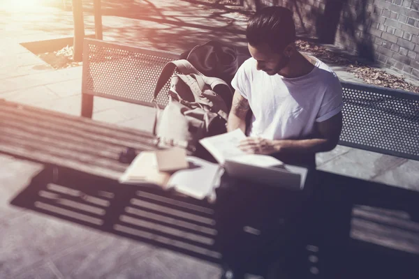 Joven vistiendo camiseta blanca sentado en el parque de la ciudad y leyendo un libro. Estudiar en la Universidad, preparación para los exámenes. Libros, cuaderno, banco de mochilas. Efecto de película horizontal, borrosa —  Fotos de Stock