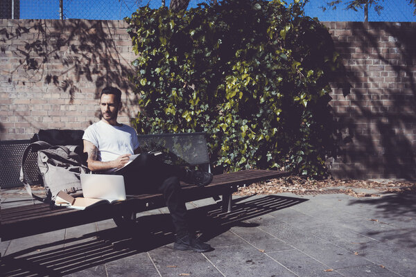 Мужчина в белой футболке сидит в городском парке и читает книгу. Обучение в университете, подготовка к экзаменам. Книги, ноутбук, скамейка для рюкзаков. Горизонтальный, эффект пленки
