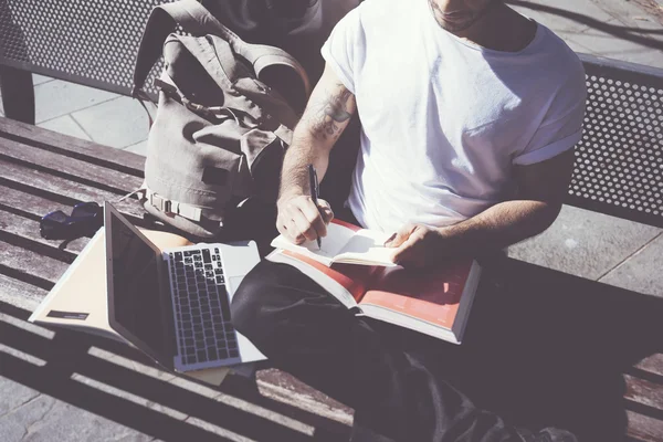 Closeup photo man wearing white tshirt sitting city park and writing notebook. Studying at the University,  preparation for exams. Books, generic design laptop, backpack bench. Horizontal, film effect