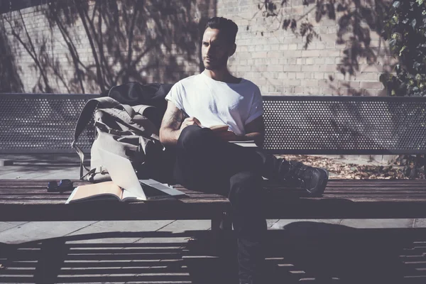 Foto homem vestindo tshirt branca sentado parque da cidade e escrevendo livro de mensagens. Estudar na Universidade, projecto de trabalho. Livros, laptop de design genérico, banco de mochila. Horizontal, efeito filme — Fotografia de Stock