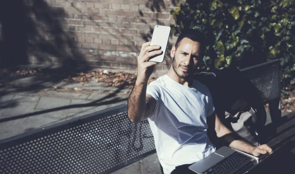 Foto homem sentado banco do parque da cidade e fazendo smartphone selfie. Usando internet sem fio. Estudando na Universidade, preparação para exames. Use livro, laptop de design genérico. Sombras horizontais e suaves — Fotografia de Stock