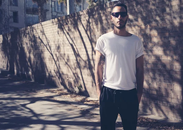 Homme barbu avec tatouage portant un t-shirt blanc vierge et des lunettes de soleil noires. Fond de mur de jardin de ville. Modélisation horizontale — Photo