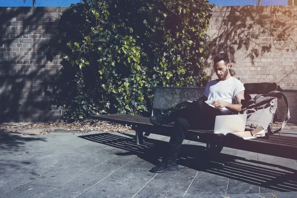 Foto barbudo homem vestindo tshirt branca sentado banco do parque da cidade e livro de leitura. A estudar na Universidade, a trabalhar num novo projecto. Livros, laptop, banco de mochilas. Horizontal — Fotografia de Stock