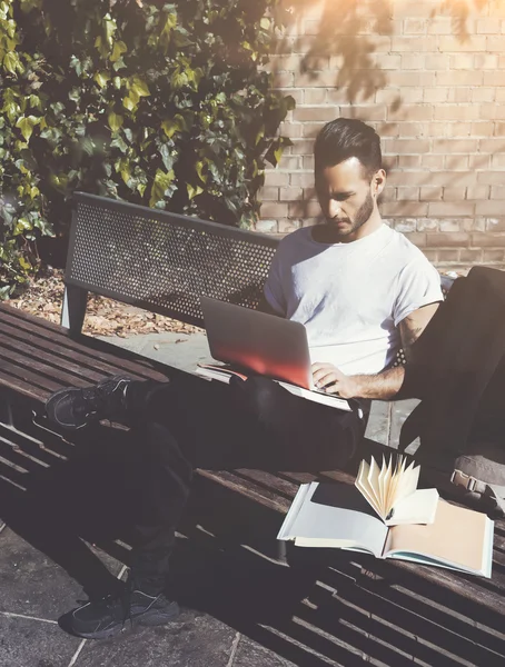 Foto uomo seduto panchina del parco della città e messaggi di testo notebook. Utilizzo di internet wireless. Studiare all'Università, preparazione agli esami. Usi il libro, il computer portatile generico di progettazione. Ombre verticali, morbide — Foto Stock