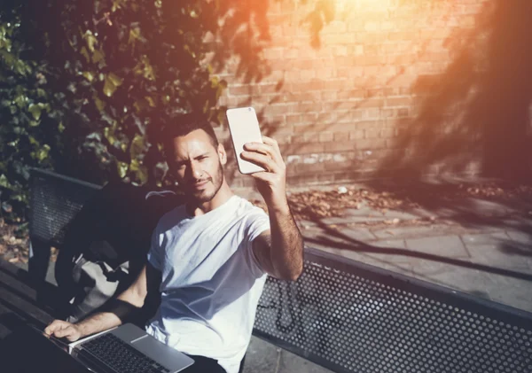 Foto homem sentado banco do parque da cidade e fazendo smartphone selfie. Usando internet sem fio. Estudando na Universidade, preparação para exames. Use livro, laptop.Horizontal design genérico, efeitos de luz solar — Fotografia de Stock