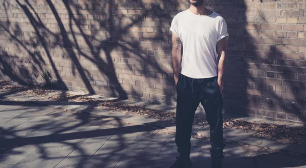 Foto homem moderno com tatuagem vestindo tshirt branca em branco. Fica em frente a uma parede de tijolos. Fundo da rua da cidade. Mockup horizontal . — Fotografia de Stock