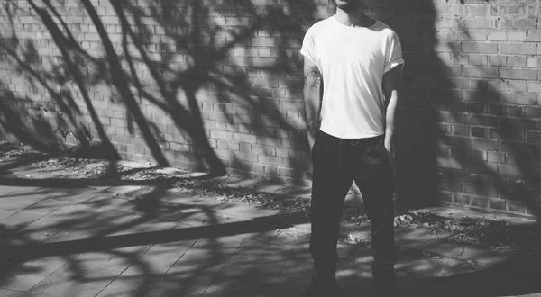 Foto homem com tatuagem vestindo tshirt branca em branco, óculos de sol. Fica em frente a uma parede de tijolos. Fundo da rua da cidade. Uma grande maquete. Preto, Branco — Fotografia de Stock