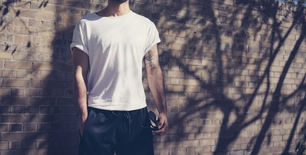 Foto estilo homem com tatuagem vestindo tshirt branca em branco. Fica em frente a uma parede de tijolos. Fundo da rua da cidade. Mockup horizontal . — Fotografia de Stock