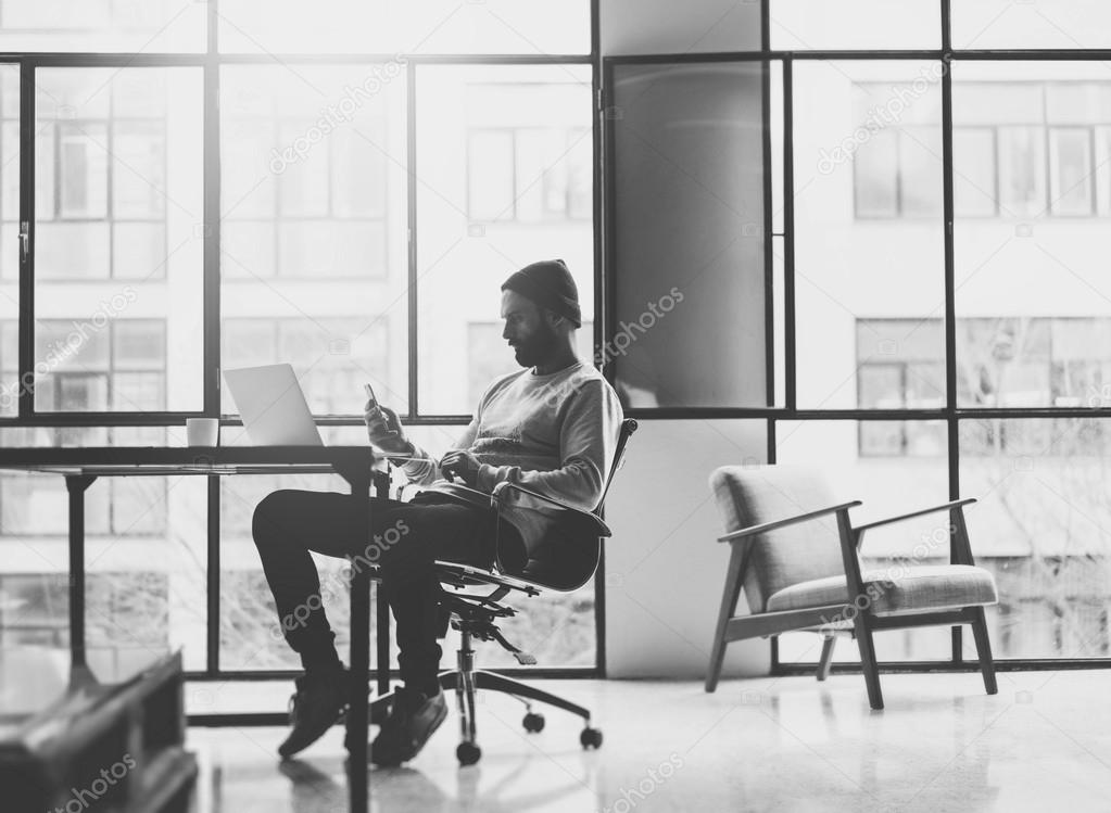 Bearded architect working on modern loft office. Texting message smartphone. Generic design notebook wood table. Horizontal mockup. Film effect. Black and white