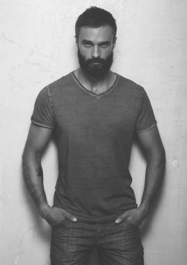 Gri boş t-shirt ve siyah kot pantolon giyen dövme ile sakallı ciddi adam, beton boş duvar karşısında duruyor. Dikey ,mockup