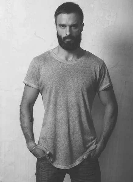 Bearded stylish man wearing gray blank t-shirt and black jeans, standing opposite empty wall. Vertical ,mockup — Stock Photo, Image