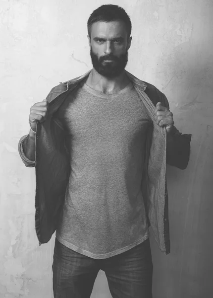 Bearded stylish man wearing gray blank shirt and black jeans, standing opposite empty wall. Vertical ,mockup — Stock Photo, Image