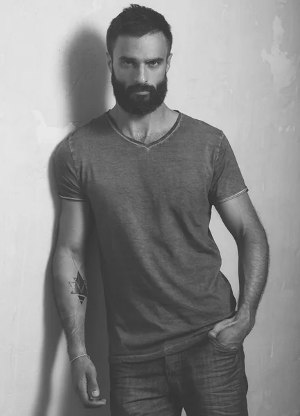 Bearded stylish man with tattoo wearing gray blank t-shirt and black jeans, standing opposite concrete empty wall. Vertical ,mockup — Stock Photo, Image