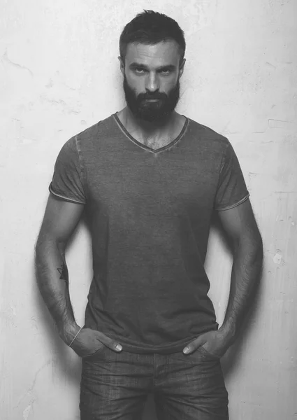 Bearded serious man with tattoo wearing gray blank t-shirt and black jeans, standing opposite concrete empty wall. Vertical ,mockup — Stock Photo, Image