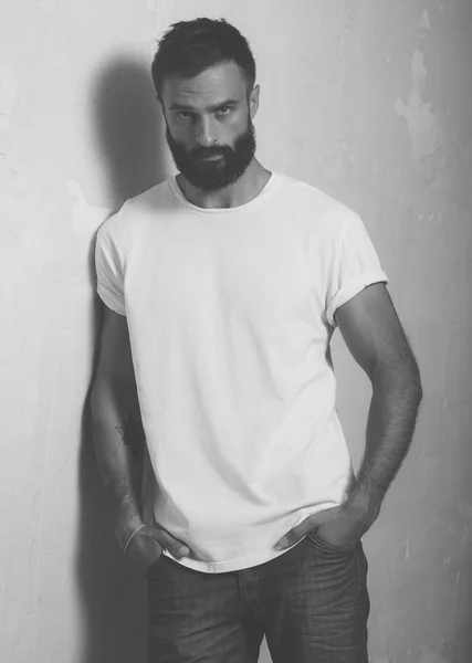 Bearded stijlvolle man met tatoeage dragen wit blanco t-shirt en zwarte jeans, staande tegenover betonnen lege muur. Verticaal, mockup — Stockfoto