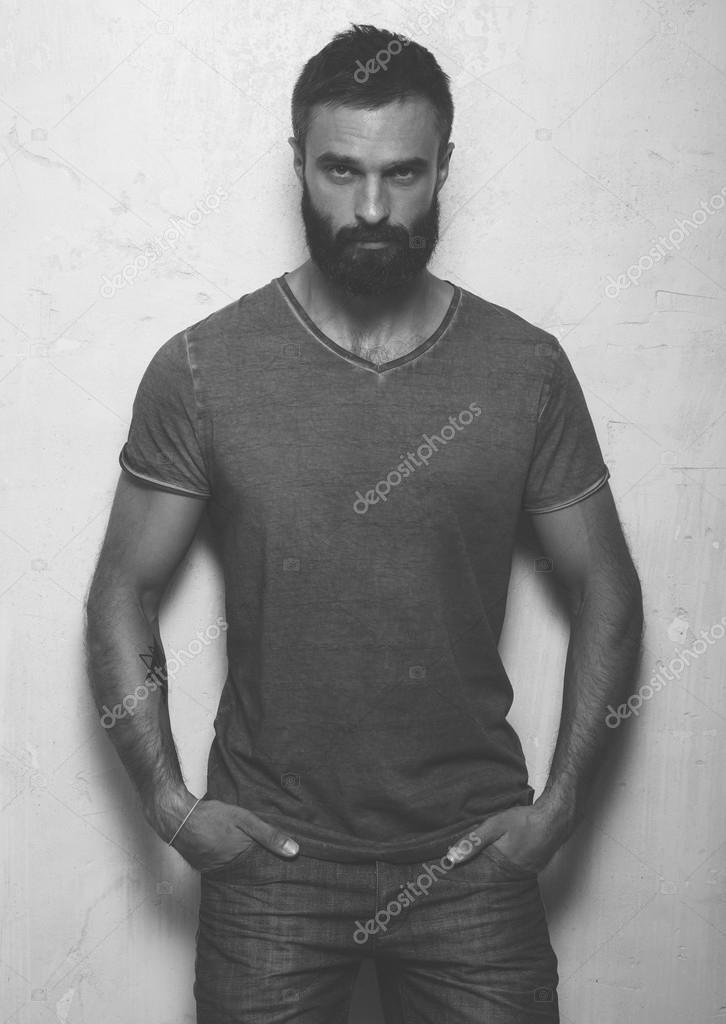 Bearded serious man with tattoo wearing gray blank t-shirt and black jeans, standing opposite concrete empty wall. Vertical ,mockup