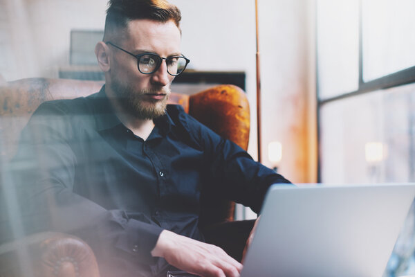 Бородатый молодой бизнесмен, работающий в современном лофт-офисе. Мужчина, сидящий в винтажном кресле. Используя современный ноутбук, панорамный фон окон. Горизонтальный, эффект пленки, размытый
