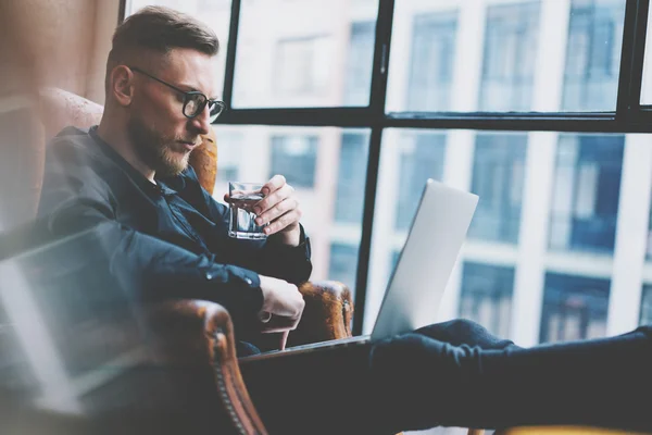 Bearded young businessman working on modern loft office. Man sitting in vintage chair, holding glass water. Using contemporary notebook, panoramic windows background. Horizontal, film effect.