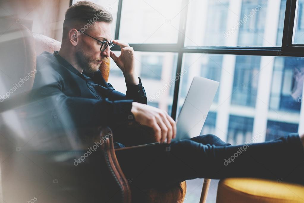 Bearded young businessman working on modern loft office. Man sitting in vintage chair. Using contemporary notebook, panoramic windows background. Horizontal, film effect.