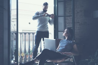 Young couple work together. Bearded man making photo smartphone beautiful girl. Working with new startup project in modern loft. Using contemporary notebook. Horizontal, film effect.
